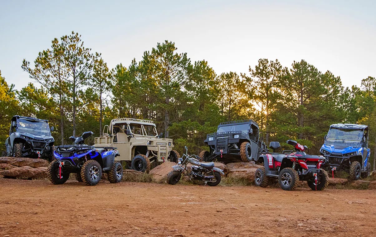 A lineup of Massimo UTVs, ATVs, and off-road vehicles on a dirt clearing among pine trees at sunset